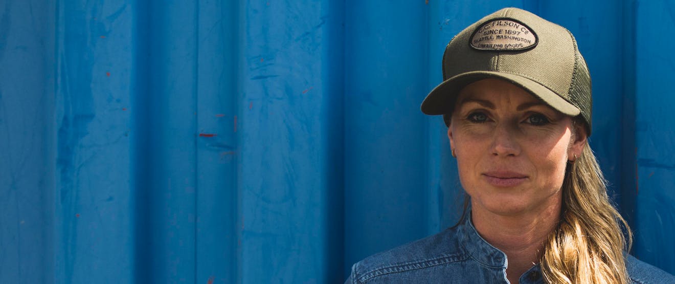 blonde Caucasian woman standing in front of a blue sided building wearing a blue button up shirt and green ball cap with the filson logo