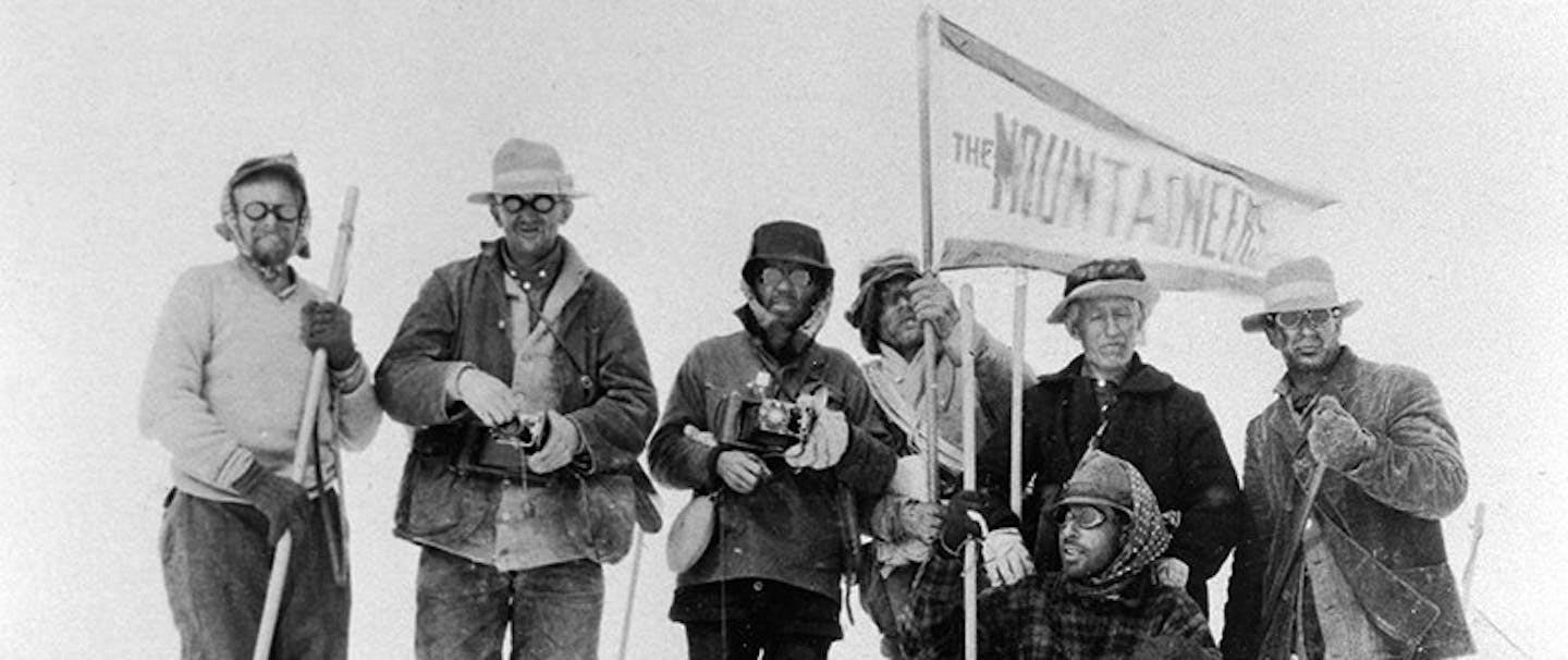 a black and white image of six men wearing a variety of jackets, hats and pants holding a flag reading the mountaineers