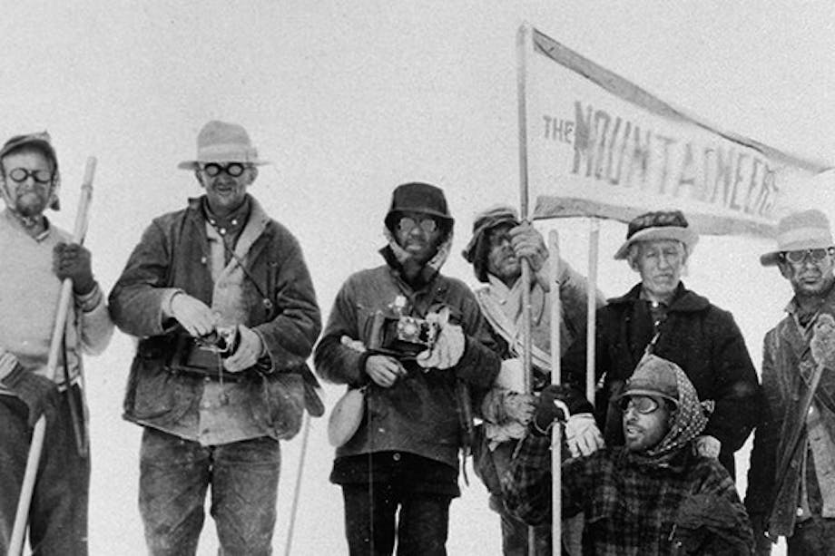 a black and white image of six men wearing a variety of jackets, hats and pants holding a flag reading the mountaineers