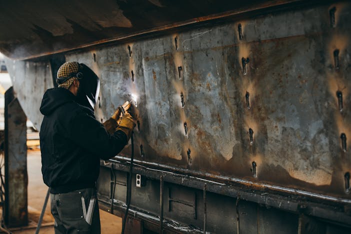 a side view of a welder wearing a brown hat, black hoodie and dark jeans welding a large piece of metal in front of him in a shop