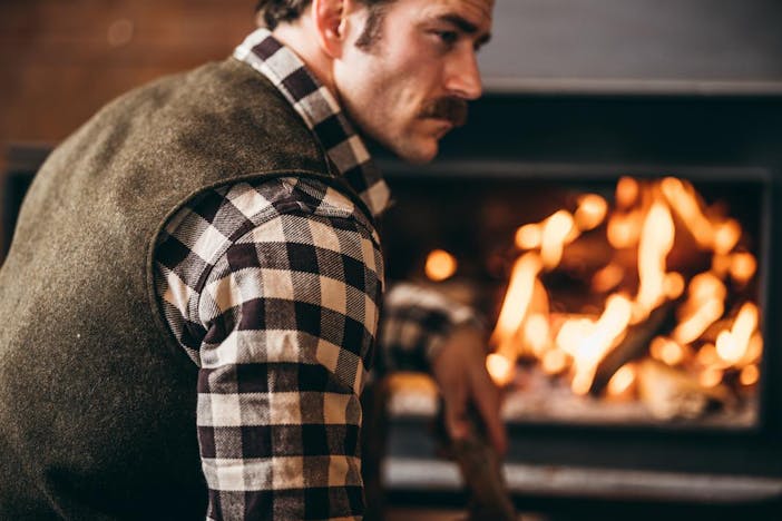 man with mustache sitting by an indoor wool fire wearing a black and white plaid shirt and green wool vest