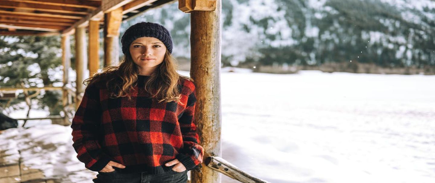 brunette woman standing on the porch of a cabin during a snowy winter wearing a red buffalo plaid sweater and hand knit beanie