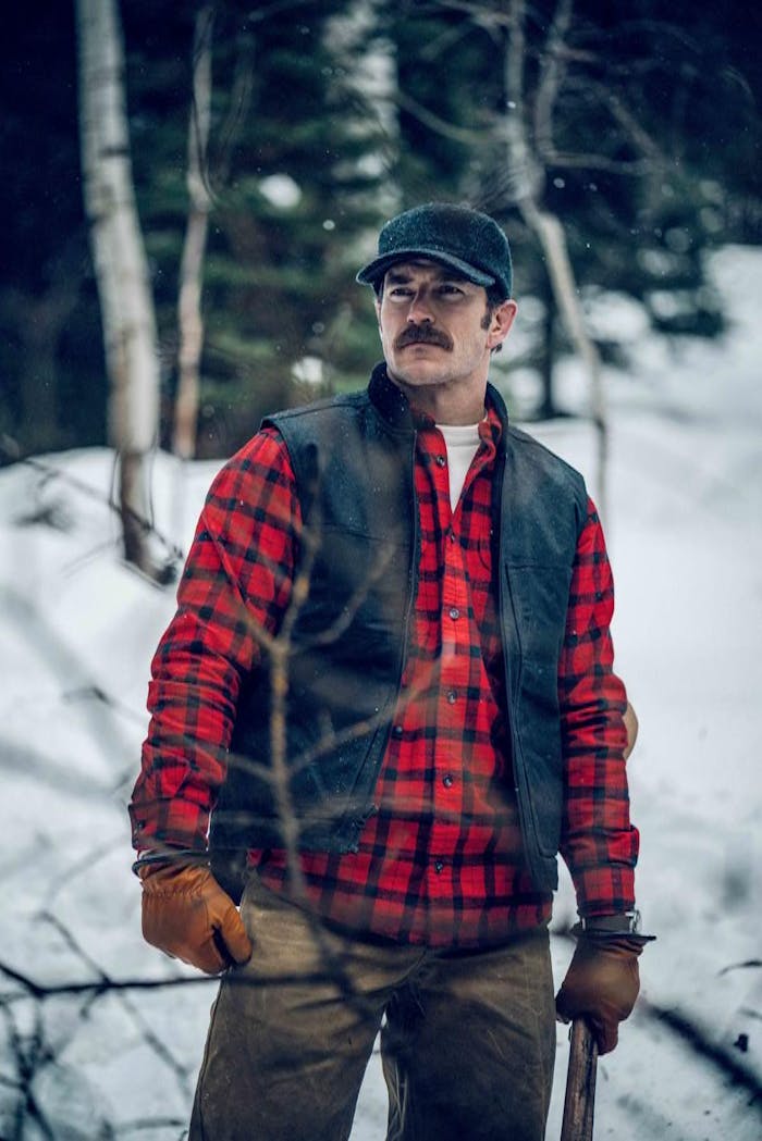 man with mustache wearing a red and black plaid flannel, grey vest, grey cap and tan gloves standing on a trail in the woods