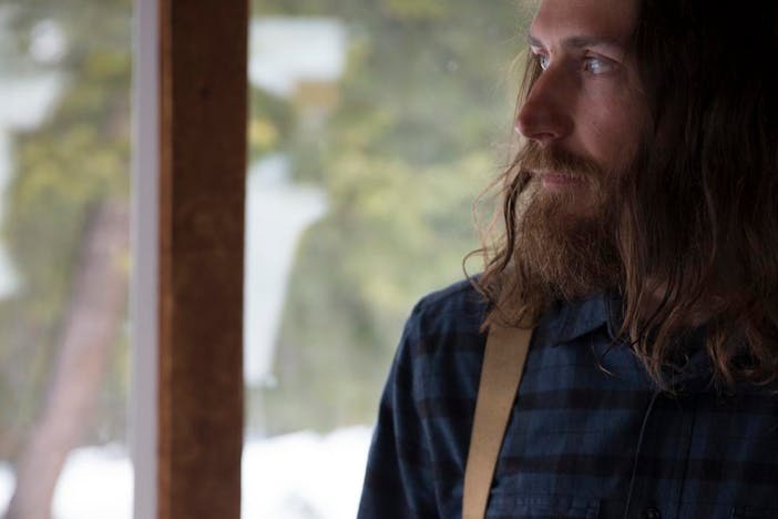 long hair and bearded man wearing a blue and black flannel with suspenders looking out a window in a cabin