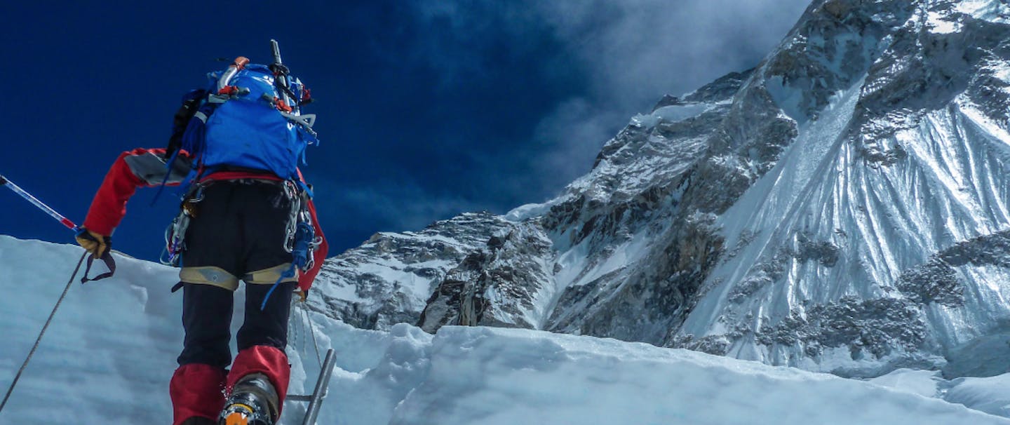 a view from behind a mounaineer scaling a large mound of ice with a metal ladder wearing black and red snow gear and a blue backpack