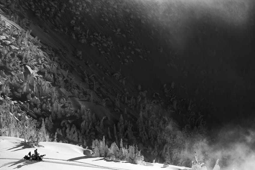 a black and white areal view of two snowmobilers looking off over the rolling snow covered mountains