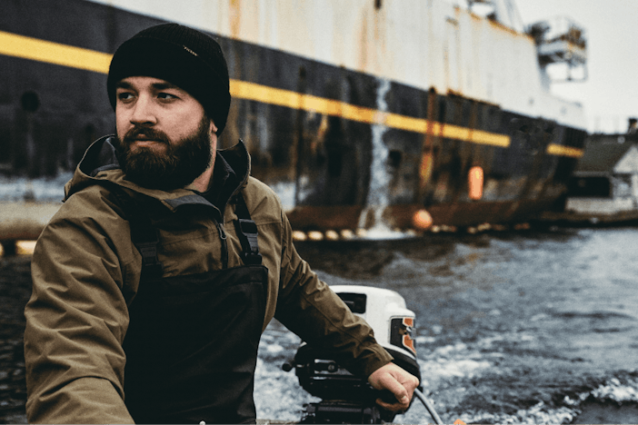 Man with black filson beanie and olive coat using hand operated motor on a boat pulling away from a larger ship