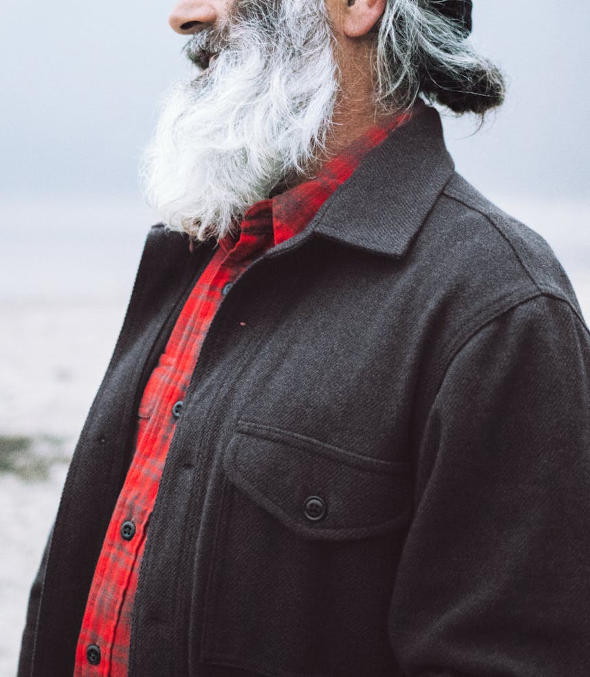 a side view of a grey and white haired and bearded man wearing a red and brown flannel shirt under a black wool button up jacket