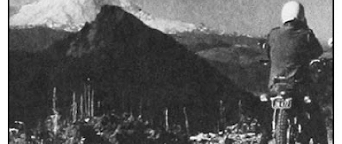 Grainy black and white image of man in white helmet and black coat looking out at large snow capped Mt. Rainier