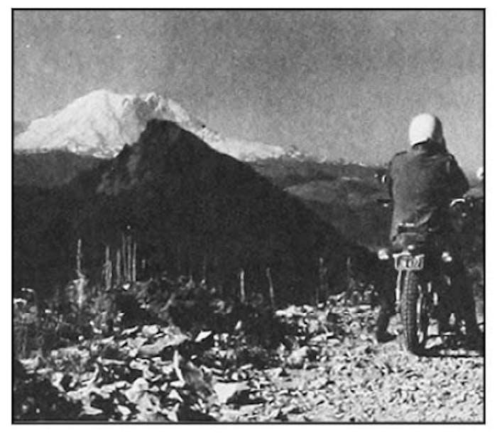 Grainy black and white image of man in white helmet and black coat looking out at large snow capped Mt. Rainier