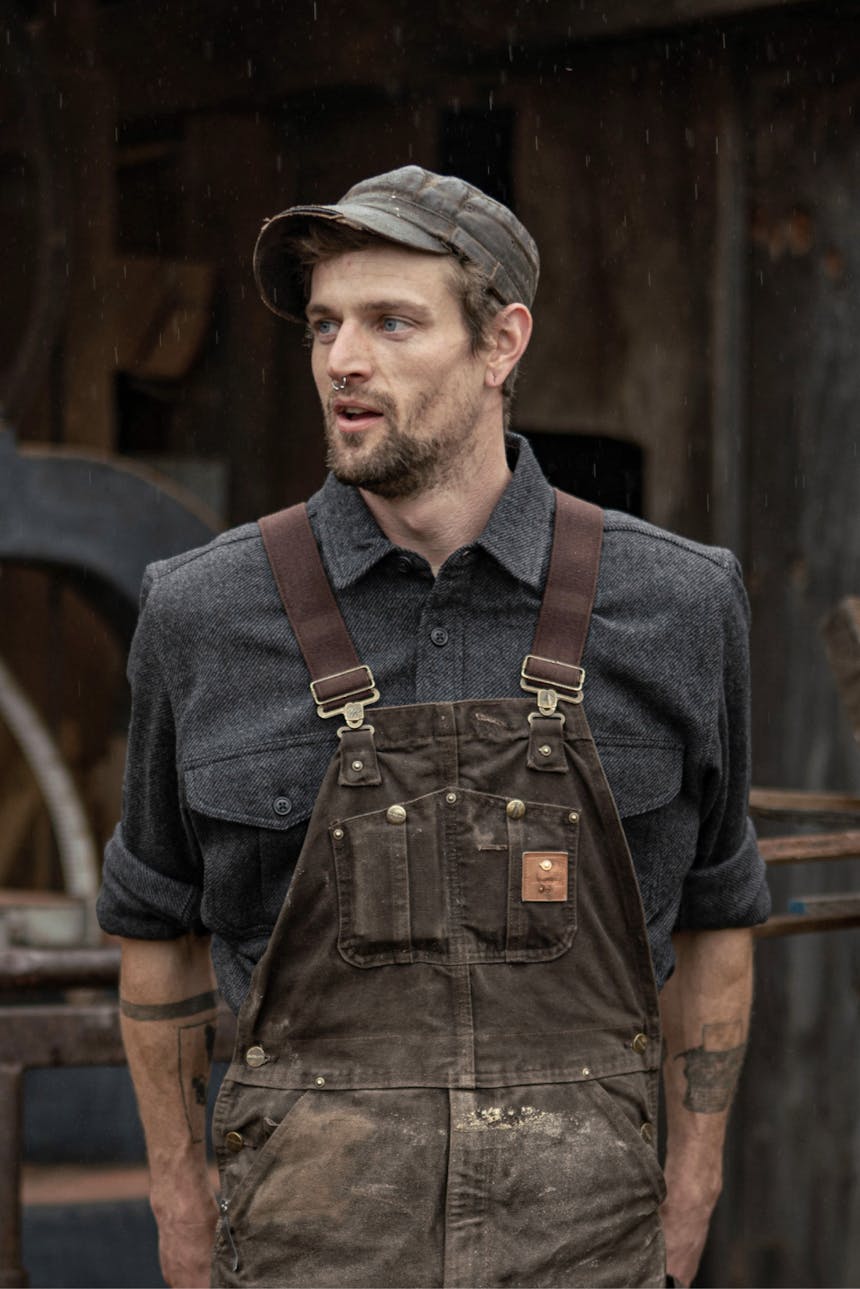 man in brown leather cap, blue buttonup shirt, and green overalls standing in front of a wooden wal