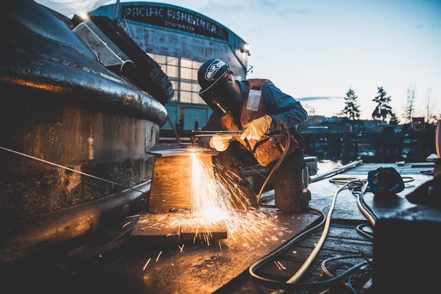 person in seahawks helmet with faceshield performing plasma cutting on piece of metal outside of pacific fisherman shipyard building