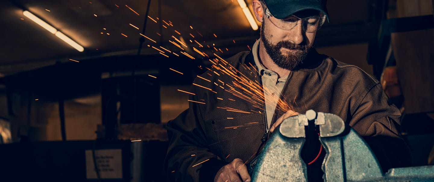man grinding metal piece held in a vice wearing a brown coat and safety glasses