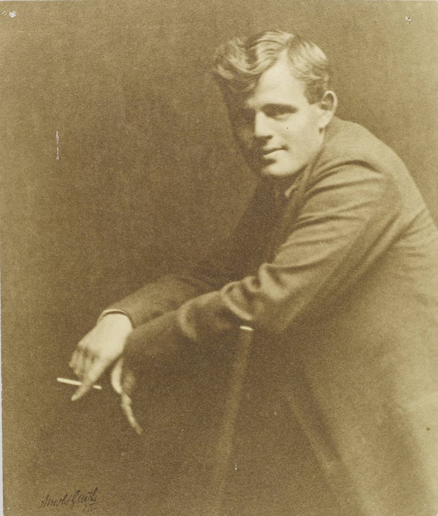 Portrait of Jack London from side, leaning over back of chair with cigarette in hand