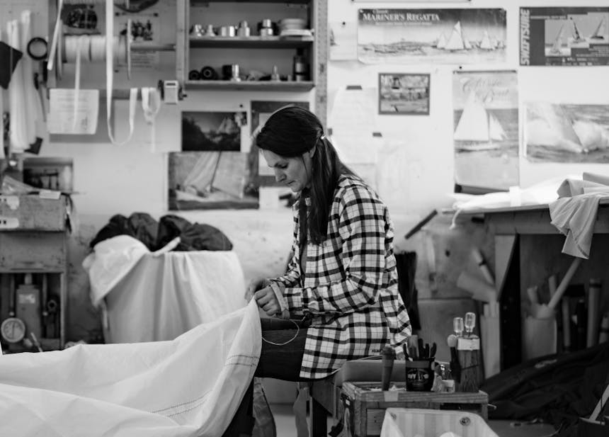 black and white image of woman in plaid shirt repairing a sail with thread and needle in a studio