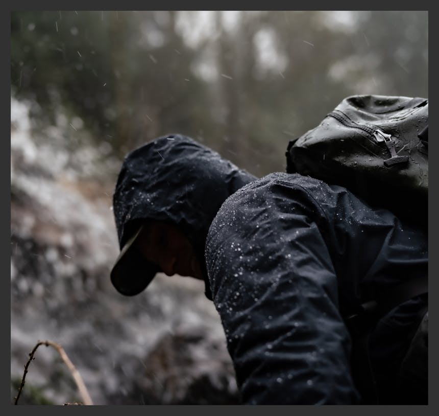 man wearing a black waterproof jacket and olive colored water proof backpack with water beading off it in a wet forest area