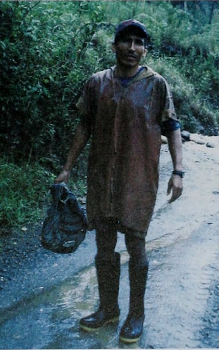 person in tan knee length garment holding a bag in his hand with galoshes on standing on a dirt road soaked in water