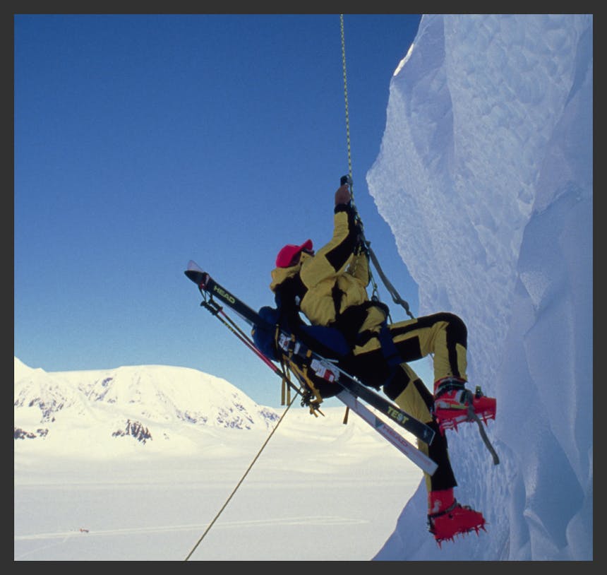 man in yellow and black skiwear and red ice climbing/skiing hybrid boots scaling a sheer ice wall with skis on his back