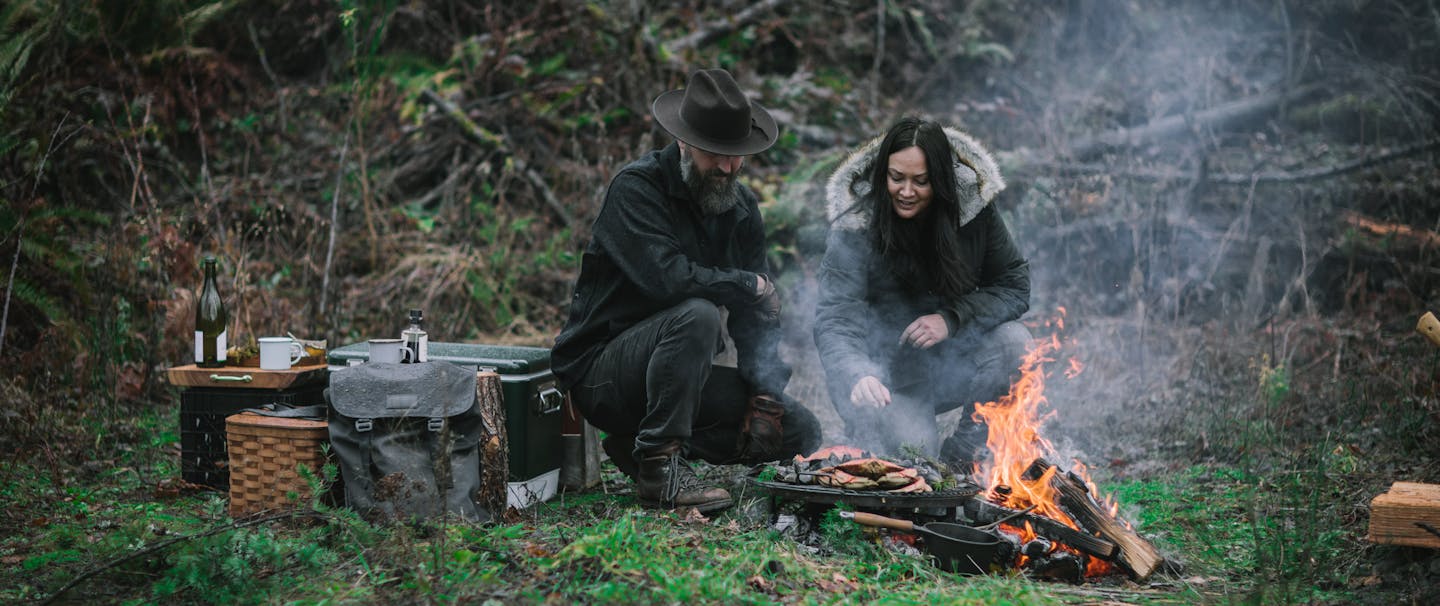 two people in lush mossy forested area tending to food cooking by an open fire