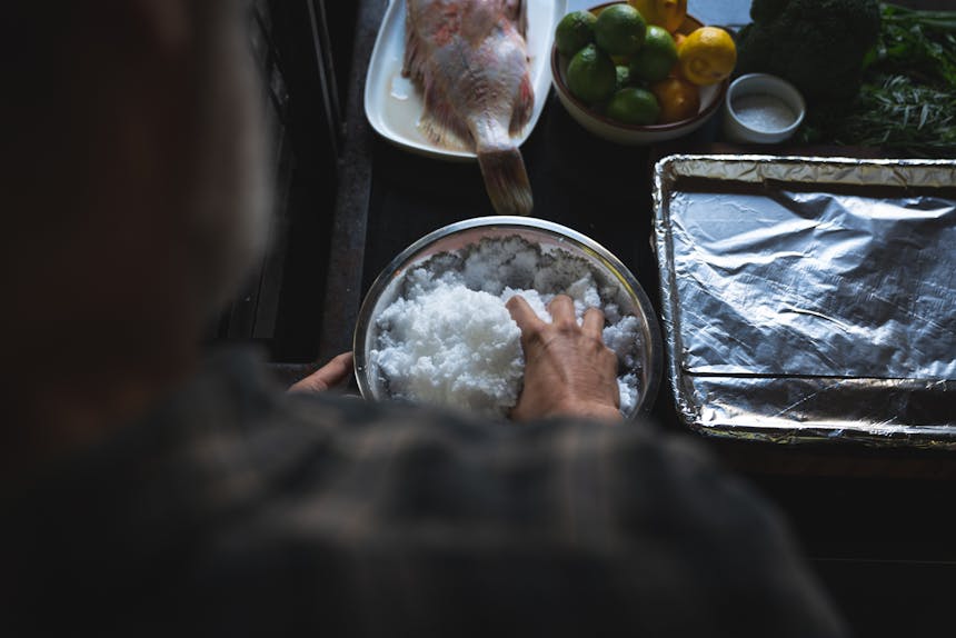 over the shoulder view of hand in bowl of salt with large fish, bowl of citrus and aluminum lined baking sheet on countertop