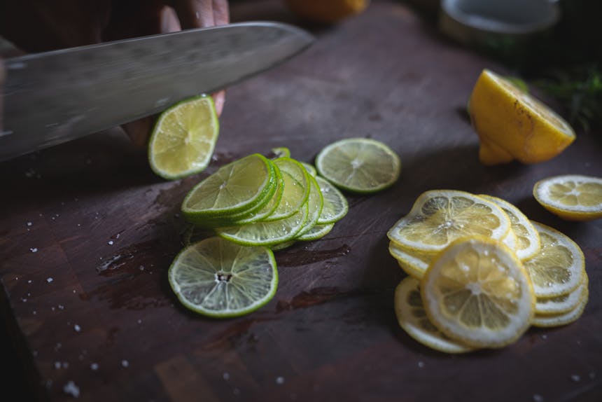 hand with knife preparing thinly sliced lime and lemon on wooden cutting board