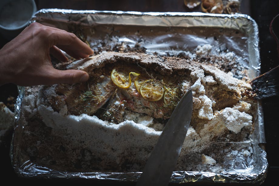 hand removing salt crust from a baked fish with lemon and herbs exposed on top of the fish skin