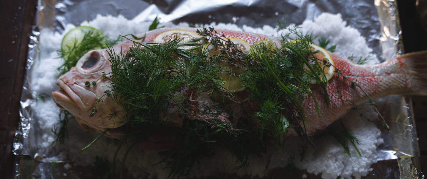 herbs and lemon wedges on top of reddish fish on bed of salt on baking sheet