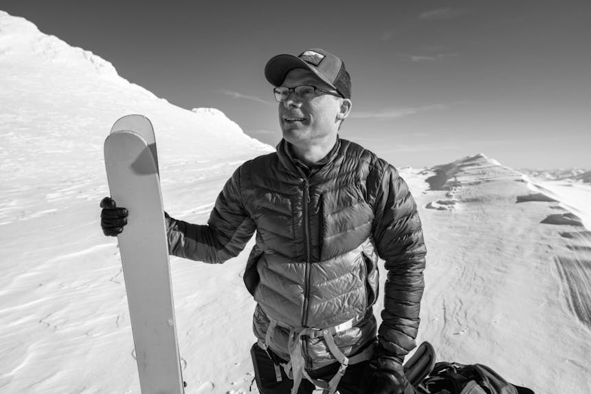 man in filson hat and puffy coat with harness on holding skis while standing on pure white snowy mountain peak