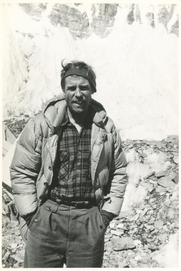 black and white portrait of man wearing a puffy coat standing in a rocky field with a large sheer snowy cliff in the background