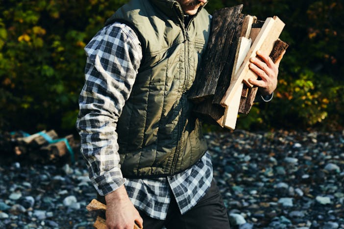 man in plaid shirt and green vest holding an armful of firewood walking on a rocky beach