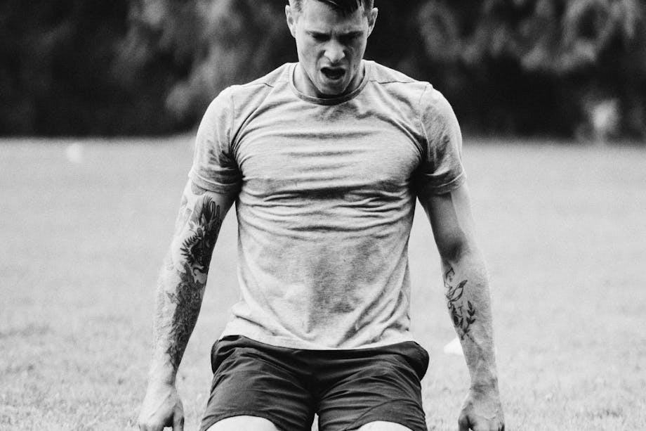 black and white image of muscular man kneeling on grass outside in gray shirt with tattoos
