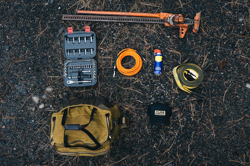 various tools for changing a tire laid out on ground with tan filson duffel and black filson hat