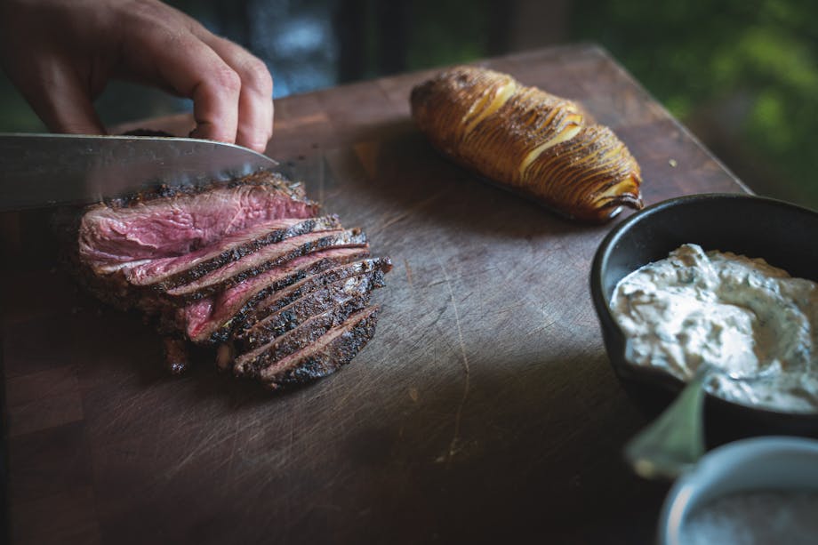 knife cutting venison sirloin held by hand next to a hasselback potato on top of a wooden cutting board with sauce in a small bowl
