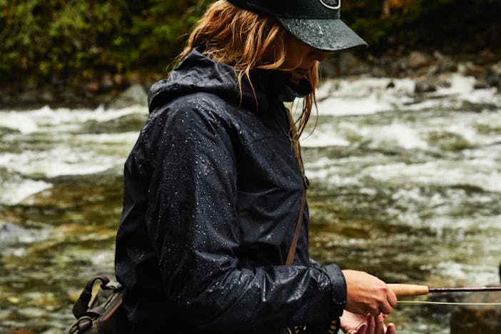 woman in waterproof black coat with water beading off of it holding a fly fishing rod standing in a river