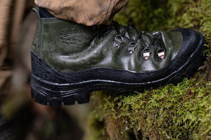 Closeup of green and black waterproof hiking boot by Danner x Filson walking over a mossy log