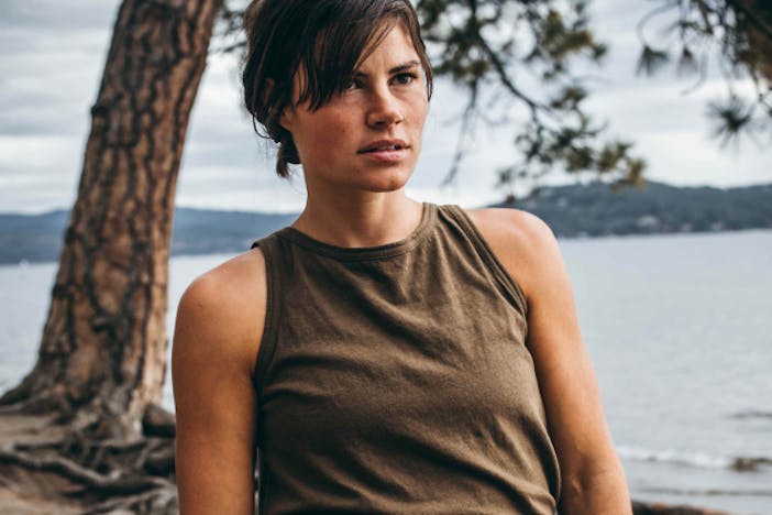 woman with short brown hair and brown tank top standing in front of a large tree growing out of the point of a beachhead at the shore of a large body of water