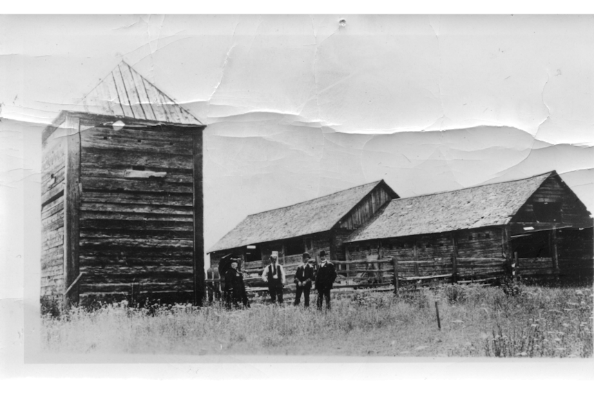 four men stand outside of ancient looking wooden prairie structures