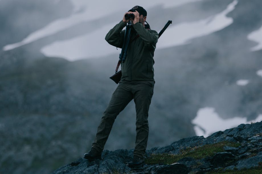 Man with rifle slung over his shoulder standing on rocky outcropping looking through field glasses wearing NeoShell Reliance Jacket