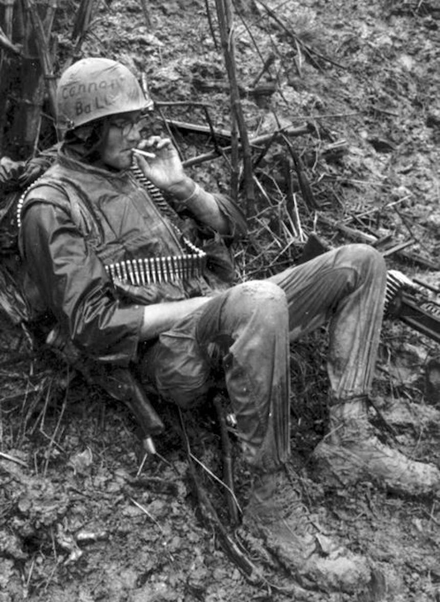 black and white image of soldier in muddy terrain in wet clothes with a bullet belt around his shoulder smoking a cigarette