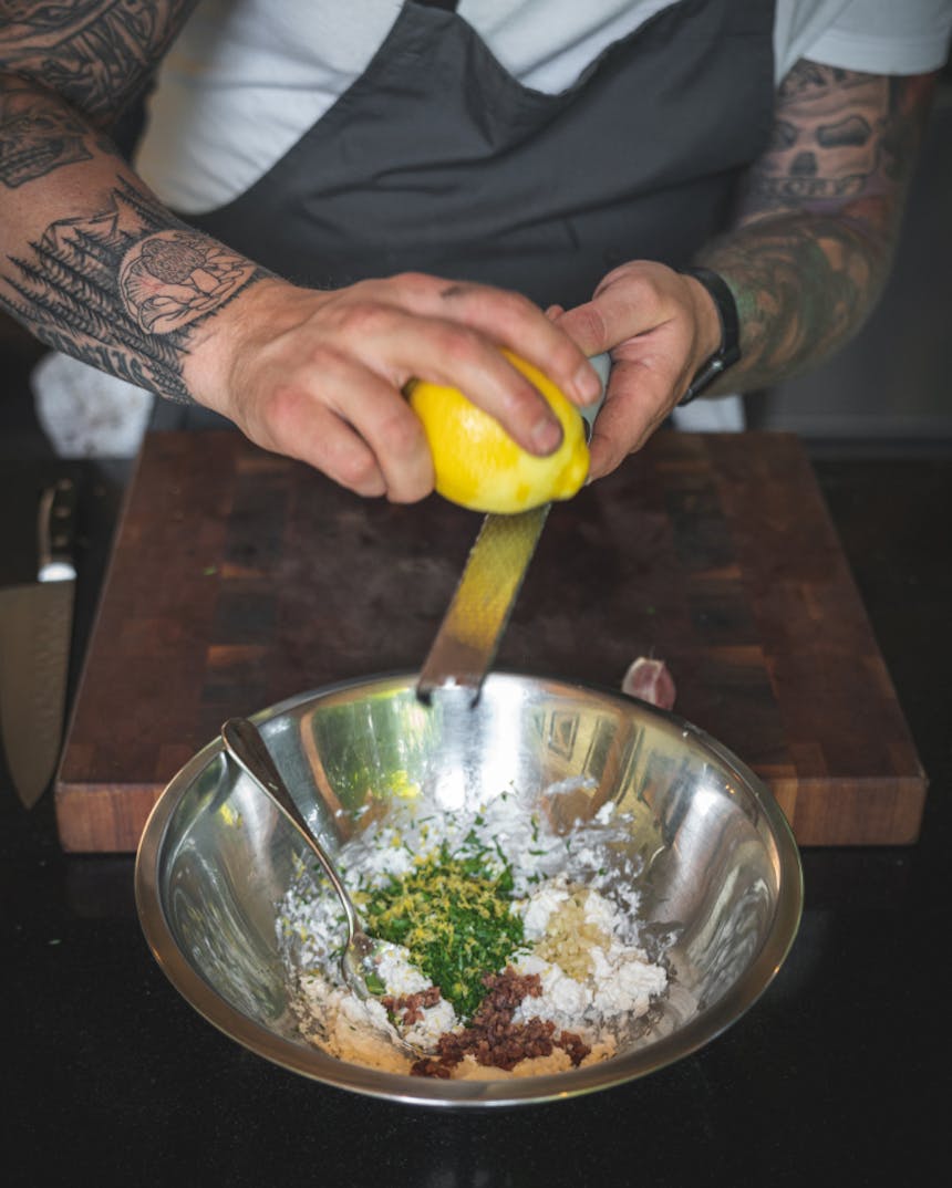 hands zesting a lemon on a long zester over a stainless steel bowl with various herbs and spices to make a sauce