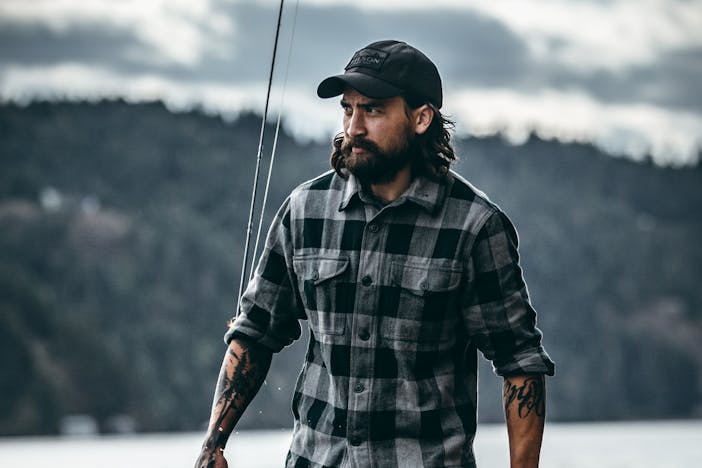 man in gray and black plaid shirt walking through ankle high water holding a fly fishing rod with pine trees in background