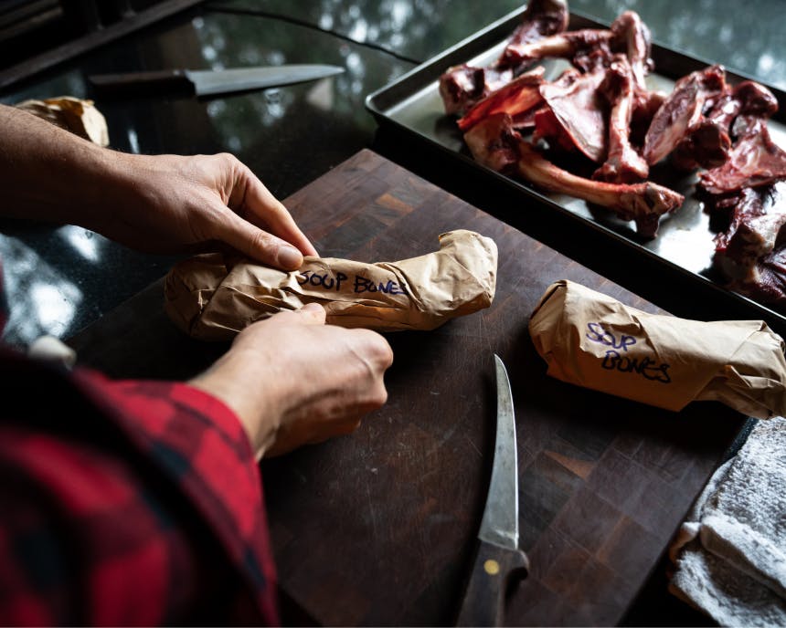 hands holding parchment wrapped soup bones with other large bones on a sheet pan behind the cutting board