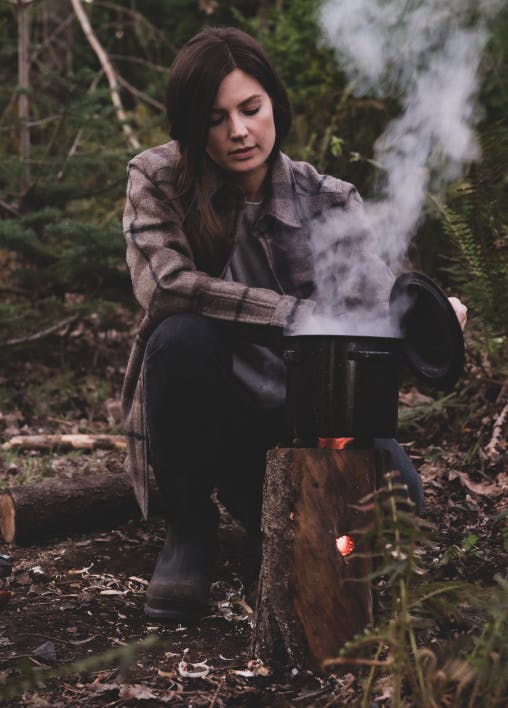 steam escaping a pot while a woman holds the lid while its sitting on top of a rocket stove log