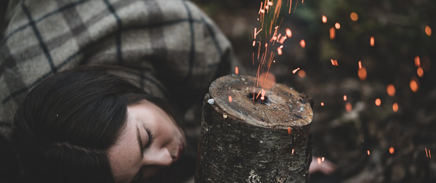 woman in brown and gray flannel shirt blowing through the side of a rocket stove sending sparks out of the central hole in the log