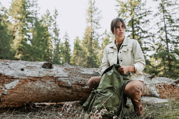 woman with white coat and green filson backpack squats in front of downed log in forest meadow