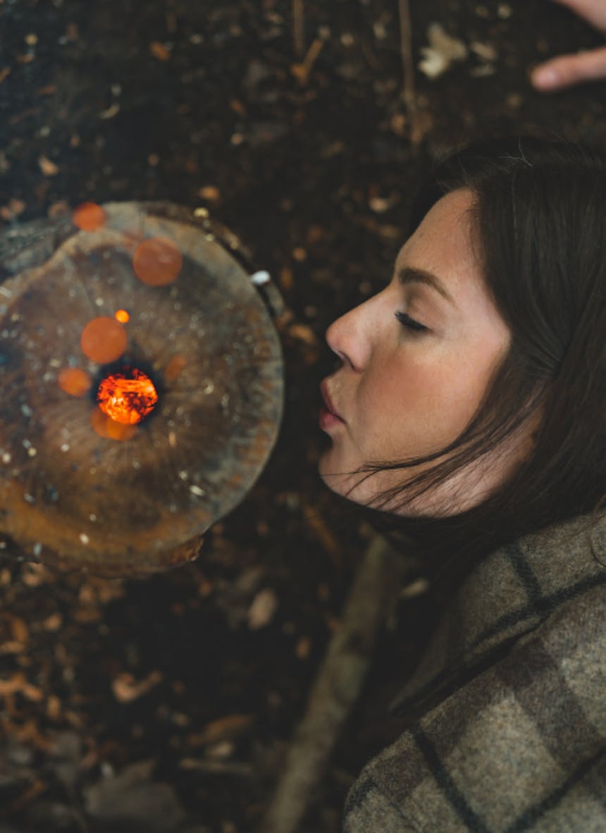 woman blowing in the side hole of a rocket stove, stoking the flames inside