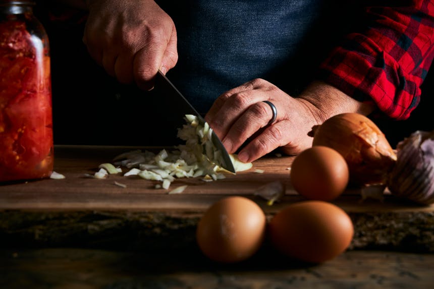 hand holding a knife chopping onions with eggs in foreground and a jar of pickled veggies