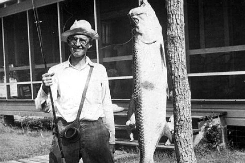 man holding fishing pole with crossbody bag and bucket hat standing next to huge fish hanging on tree