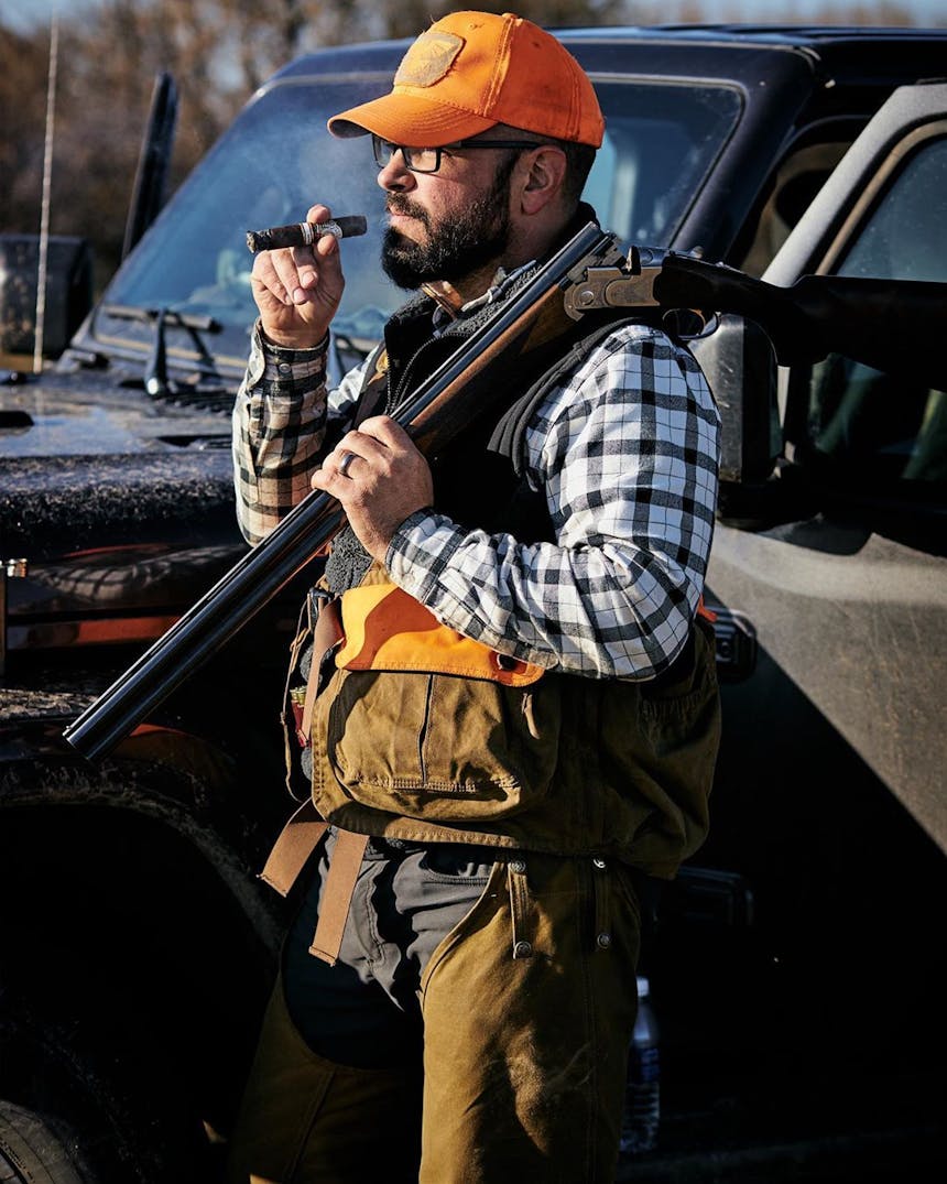 Rossano Russo smoking a cigar with a shotgun slung over his shoulder wearing hunting gear and an orange cap