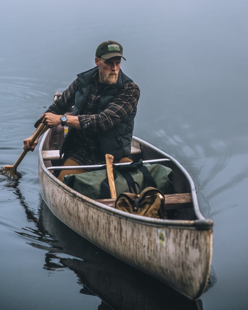 man in vest and brown filson cap paddling a single person canoe with waterproof duffel in boat